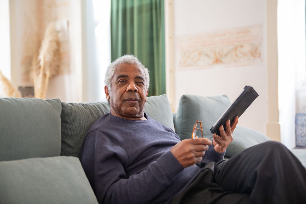 An elderly Black man relaxing on the couch at home while holding his tablet